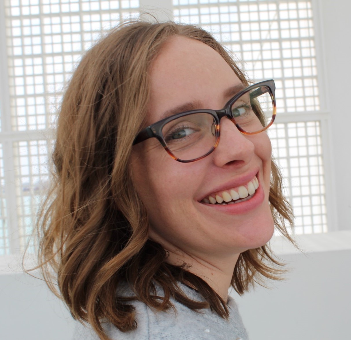 Woman with shoulder-length brown hair and wearing glasses smiles against a white background.
