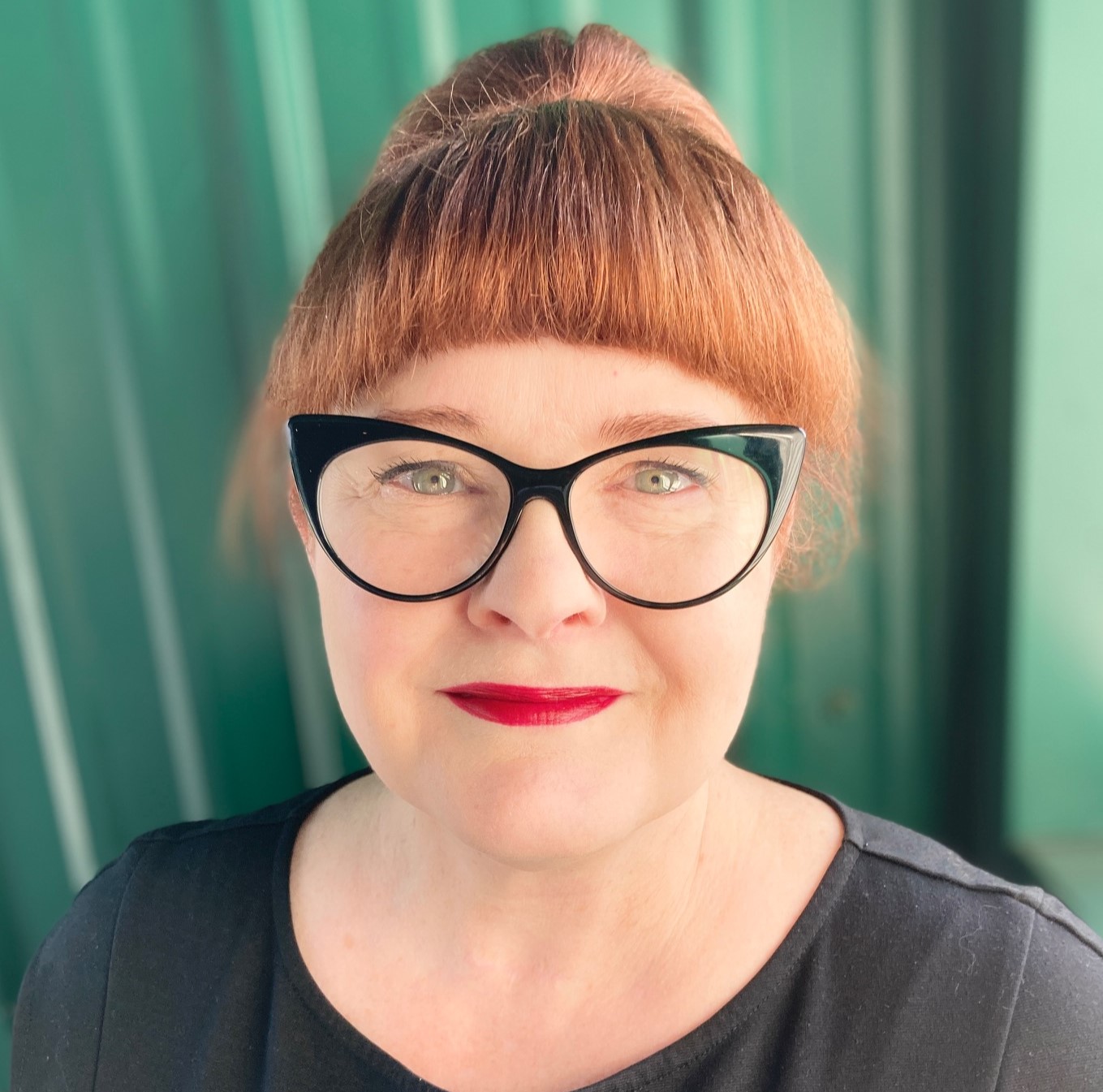 This is a headshot of Traci, a white woman with red hair tied up in a bun. She has black glasses and wears a black dress. She is standing in front of a teal-colored background.