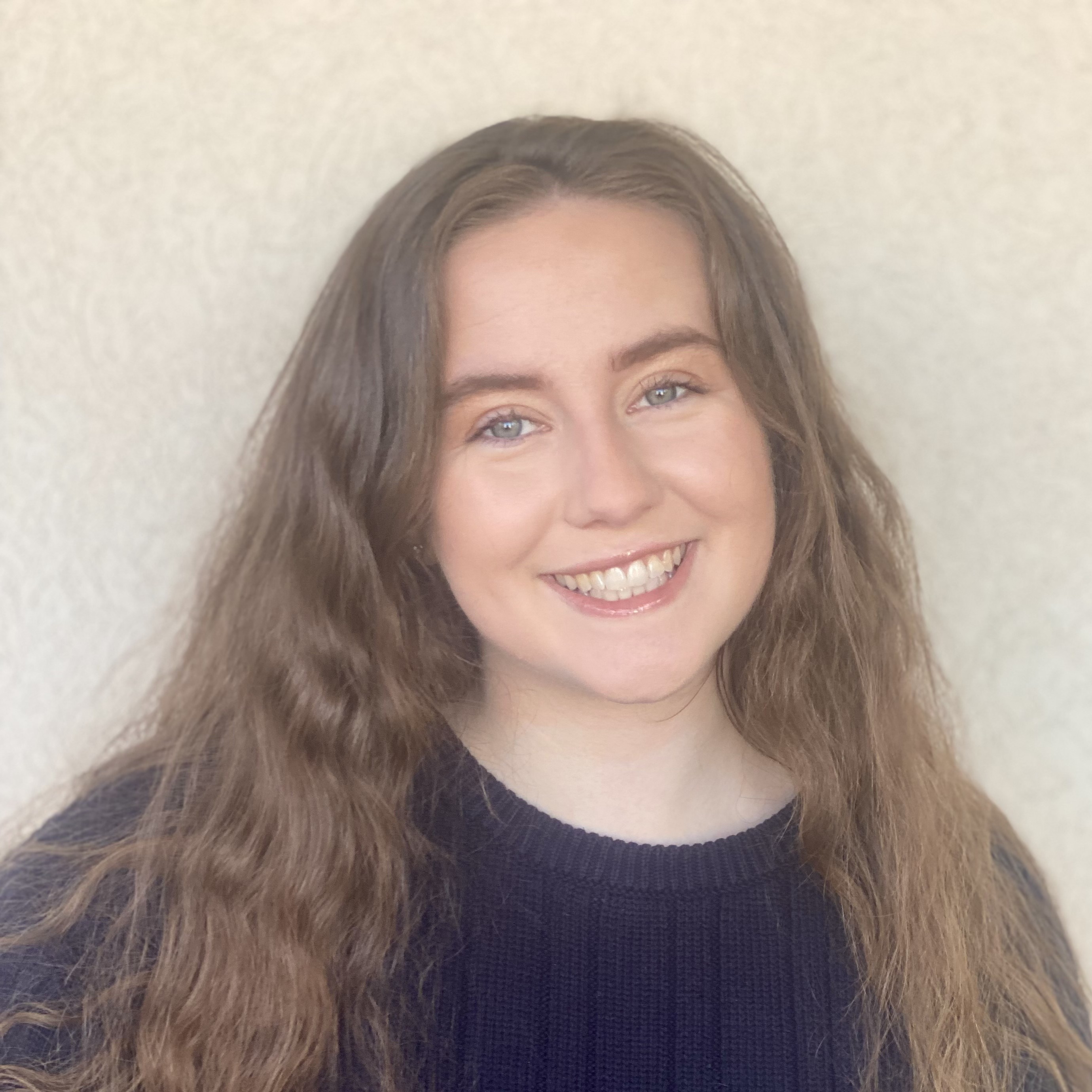 Cara Elzie is a white woman with light brown hair and gray-blue eyes. in this photo, she is wearing a navy blue sweater and is standing in front of a tan background